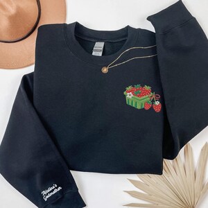May include: Black sweatshirt with an embroidered design of strawberries in a green basket. A gold necklace with a small pendant rests on the sweatshirt. The sleeve features the text "Adeline's Godmother".