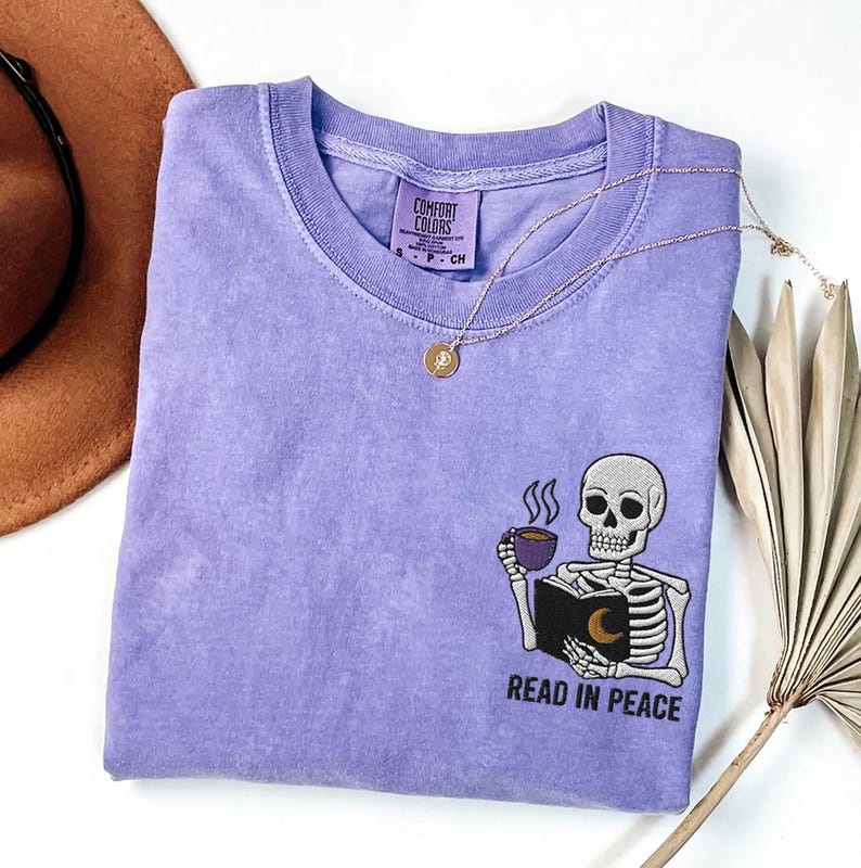 Puede incluir: Camiseta morada con un dise&ntilde;o bordado de un esqueleto sosteniendo un libro y una taza de caf&eacute;. El texto "Read in Peace" est&aacute; debajo del esqueleto.