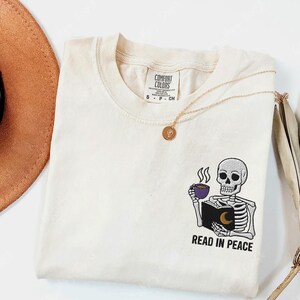 Puede incluir: Una camiseta blanca con un dise&ntilde;o bordado de un esqueleto sosteniendo un libro y una taza de caf&eacute;. El texto "Read in Peace" est&aacute; debajo del esqueleto.