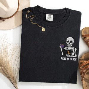 Puede incluir: Camiseta negra con un dise&ntilde;o bordado de un esqueleto sosteniendo un libro y una taza de caf&eacute;. El texto "READ IN PEACE" est&aacute; bordado debajo del esqueleto.