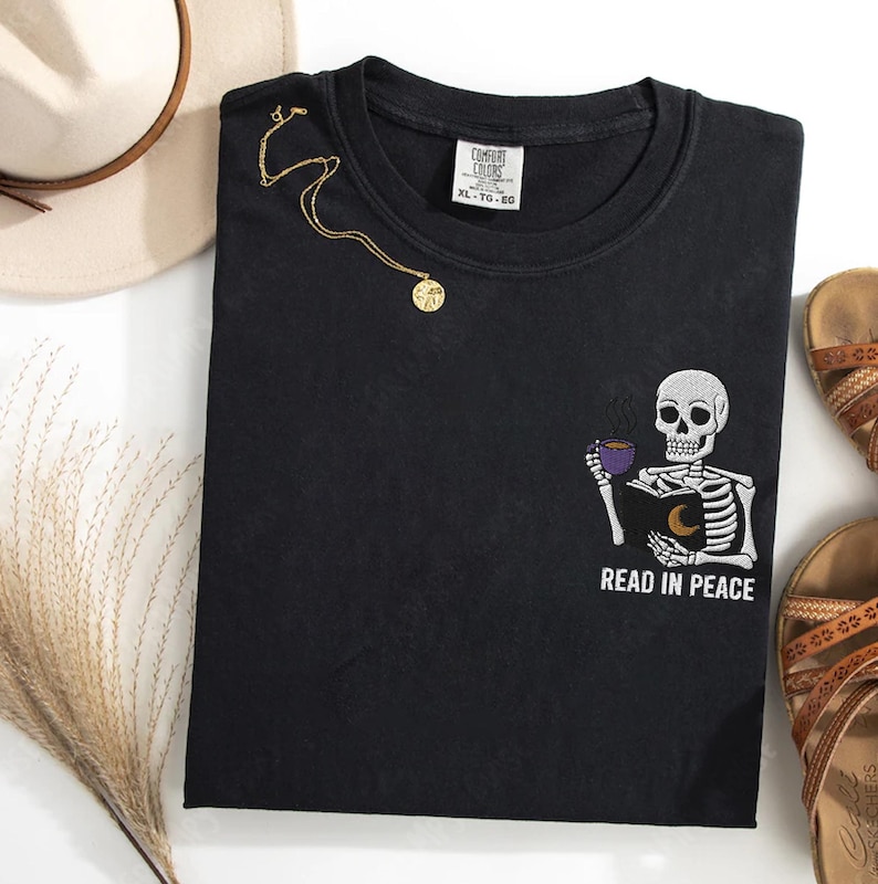 Puede incluir: Camiseta negra con un dise&ntilde;o bordado de un esqueleto sosteniendo un libro y una taza de caf&eacute;. El texto "READ IN PEACE" est&aacute; bordado debajo del esqueleto.