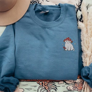 K&ouml;nnte beinhalten: Ein blaues Sweatshirt mit einem gestickten Design eines wei&szlig;en Katers, der einen roten Pilz-Hut tr&auml;gt. Die Katze h&auml;lt einen kleinen roten Pilz.