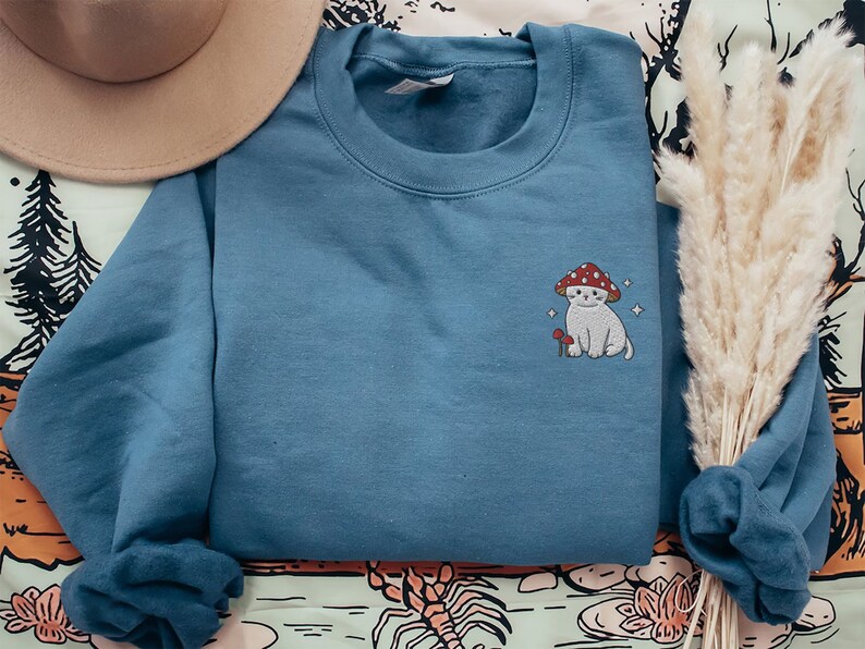 K&ouml;nnte beinhalten: Ein blaues Sweatshirt mit einem gestickten Design eines wei&szlig;en Katers, der einen roten Pilz-Hut tr&auml;gt. Die Katze h&auml;lt einen kleinen roten Pilz.