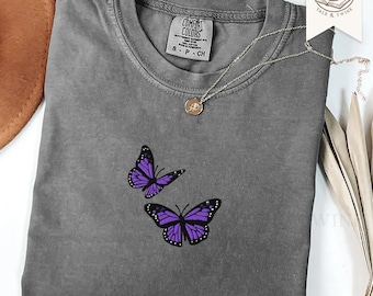 May include: A charcoal gray short-sleeve t-shirt featuring an embroidered design of two purple butterflies with black details. The shirt has a crew neckline and a Comfort Colors brand label at the inner collar. A gold-tone necklace rests on the fabric.