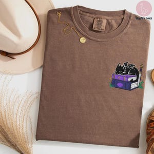 May include: Brown Comfort Colors t-shirt with a small embroidered design of a black dragon sleeping on purple books. A gold necklace with a pendant is draped on the shirt. The tag reads XL-TG-EG.
