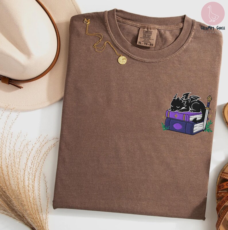 May include: Brown Comfort Colors t-shirt with a small embroidered design of a black dragon sleeping on purple books. A gold necklace with a pendant is draped on the shirt. The tag reads XL-TG-EG.