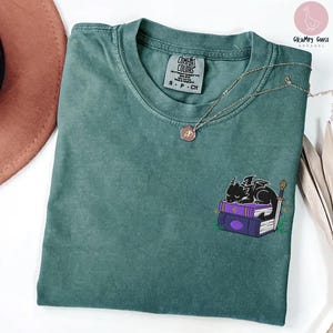 May include: A teal Comfort Colors t-shirt with a small embroidered design of a black dragon sleeping on purple books. A silver necklace with a pendant is on the shirt. The shirt has a tag that says "Comfort Colors".