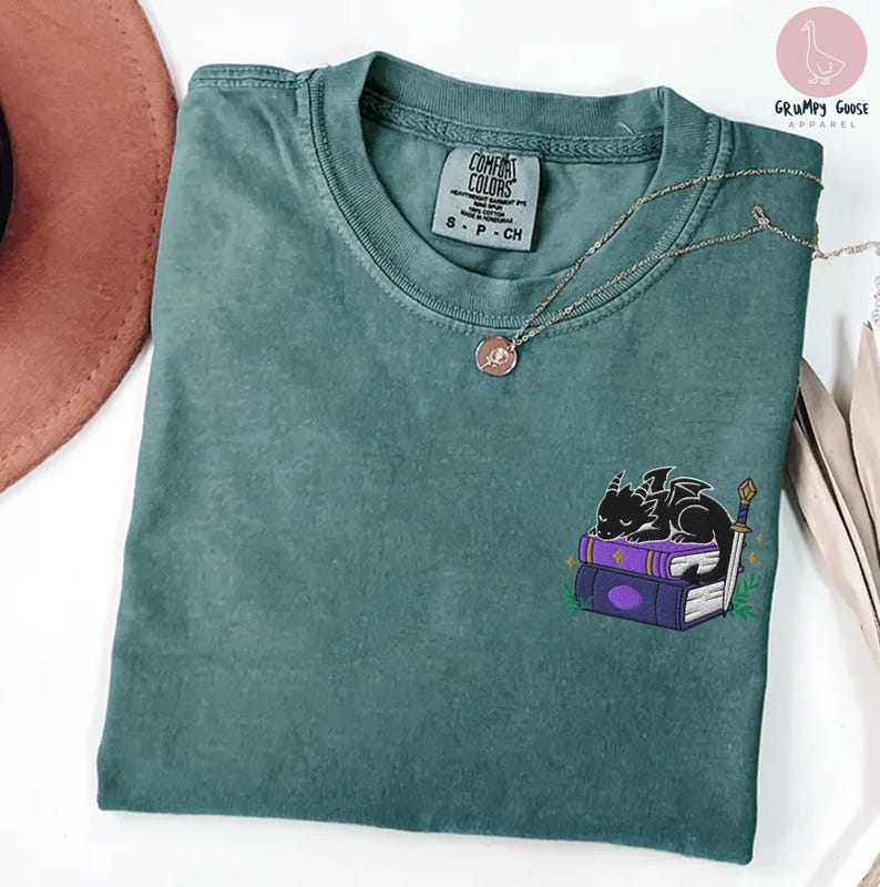 May include: A teal Comfort Colors t-shirt with a small embroidered design of a black dragon sleeping on purple books. A silver necklace with a pendant is on the shirt. The shirt has a tag that says "Comfort Colors".