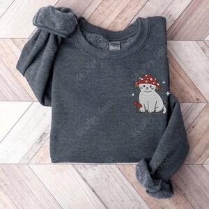 K&ouml;nnte beinhalten: Ein dunkelgrauer Pullover mit einer wei&szlig;en Katze, die einen roten Pilz-Hut tr&auml;gt, der auf der Vorderseite gestickt ist. Die Katze sitzt neben einem kleinen Pilz.