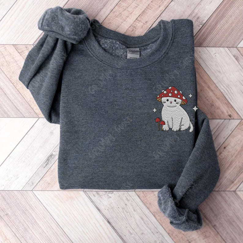 K&ouml;nnte beinhalten: Ein dunkelgrauer Pullover mit einer wei&szlig;en Katze, die einen roten Pilz-Hut tr&auml;gt, der auf der Vorderseite gestickt ist. Die Katze sitzt neben einem kleinen Pilz.