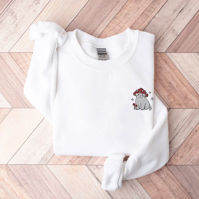 K&ouml;nnte beinhalten: Wei&szlig;er Pullover mit einem kleinen gestickten Design eines Katers, der einen roten Pilz auf dem Kopf tr&auml;gt. Der Kater sitzt neben einem kleinen Pilz.