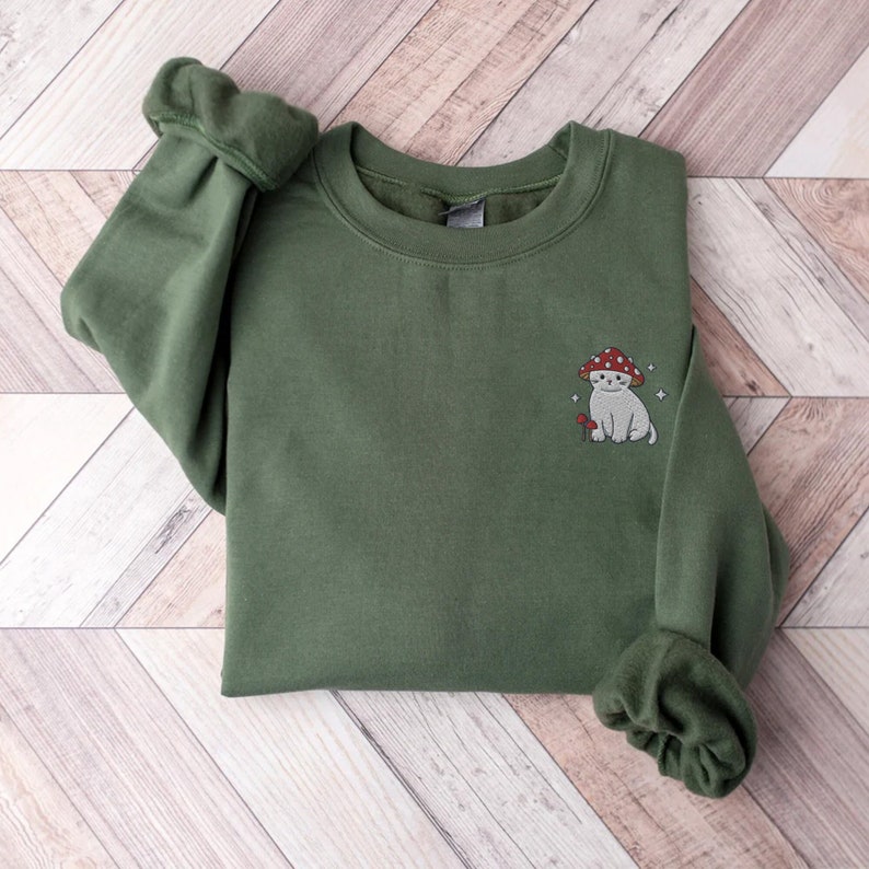 K&ouml;nnte beinhalten: Ein gr&uuml;ner Pullover mit einem gestickten Motiv eines wei&szlig;en Katers, der einen roten Pilz-Hut tr&auml;gt. Die Katze h&auml;lt einen kleinen Pilz in ihrer Pfote.