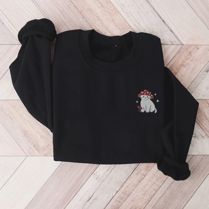 K&ouml;nnte beinhalten: Schwarzer Pullover mit einem wei&szlig;en K&auml;tzchen, das einen roten Pilz-Hut tr&auml;gt, auf der Vorderseite gestickt. Die Katze befindet sich neben einem kleinen Pilz und ist von kleinen wei&szlig;en Sternen umgeben.