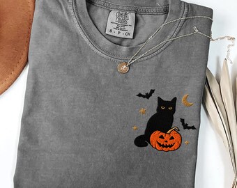 May include: A charcoal gray short-sleeve t-shirt featuring an embroidered design on the chest. The embroidery depicts a black cat sitting on an orange jack-o'-lantern, surrounded by two small black bats, a crescent moon, and two gold stars.