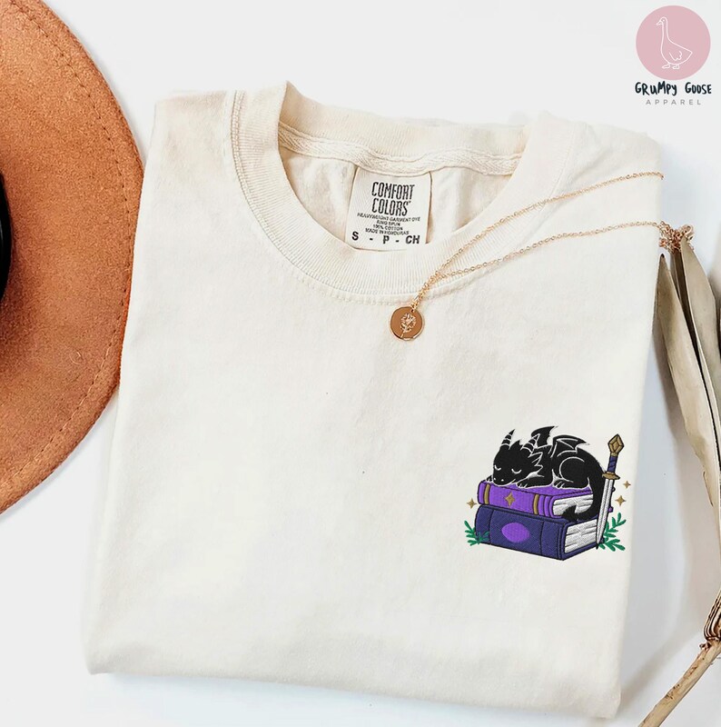 May include: Cream-colored Comfort Colors t-shirt with a small embroidered design of a black dragon sleeping on purple books with a sword. A gold necklace and a brown hat are also visible.