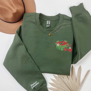 May include: An olive green sweatshirt featuring an embroidered strawberry basket design. A gold necklace with a pendant is draped on the sweatshirt. The sleeve is embroidered with the text "Adeline's Godmother". A brown hat is visible in the background.