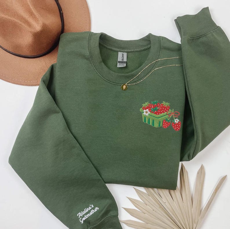May include: An olive green sweatshirt featuring an embroidered strawberry basket design. A gold necklace with a pendant is draped on the sweatshirt. The sleeve is embroidered with the text "Adeline's Godmother". A brown hat is visible in the background.