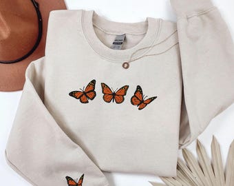 May include: A beige crewneck sweatshirt featuring three embroidered monarch butterflies across the chest and one smaller monarch butterfly embroidered on the left sleeve cuff. The garment has a ribbed collar, cuffs, and hem.