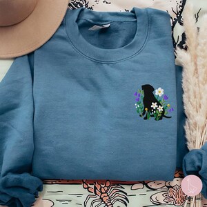 Pu&ograve; includere: Una felpa blu con un ricamo di un labrador nero circondato da fiori bianchi e viola. La felpa ha un girocollo e maniche lunghe. Un cappello beige &egrave; visibile in alto a sinistra.