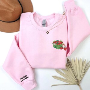 May include: A pale pink sweatshirt featuring an embroidered strawberry basket design. The sleeve is embroidered with the text "Adeline's Godmother". A gold necklace and a brown hat are also visible.