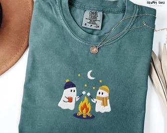 May include: A teal crewneck t-shirt featuring an embroidered design of two ghosts roasting marshmallows over a campfire under a crescent moon and stars. The shirt has a Comfort Colors label at the neck and a thin gold-toned chain necklace.