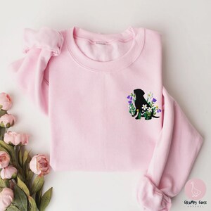 Pu&ograve; includere: Felpa rosa chiaro con una silhouette nera di cane circondata da fiori ricamati viola, bianchi e verdi. La felpa ha un girocollo e maniche lunghe. Il logo Grumpy Goose Apparel si trova in basso a destra.