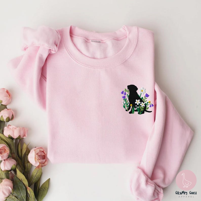Pu&ograve; includere: Felpa rosa chiaro con una silhouette nera di cane circondata da fiori ricamati viola, bianchi e verdi. La felpa ha un girocollo e maniche lunghe. Il logo Grumpy Goose Apparel si trova in basso a destra.