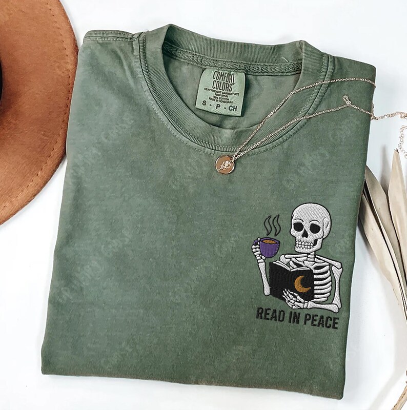 Puede incluir: Una camiseta verde con un esqueleto leyendo un libro y sosteniendo una taza de caf&eacute;. El texto "Read in Peace" est&aacute; bordado debajo del esqueleto.