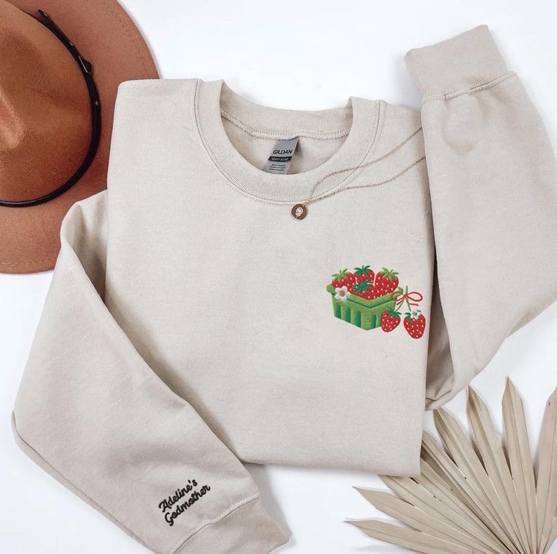 May include: Beige crew neck sweatshirt featuring an embroidered strawberry basket design. A gold necklace and the text "Adeline's Godmother" on the sleeve are visible. The sweatshirt is made of a soft material.