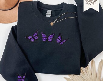 May include: A black crewneck sweatshirt featuring three embroidered purple butterflies across the chest and one smaller purple butterfly embroidered on the left sleeve cuff. The garment has a Gildan brand label at the inner neckline.