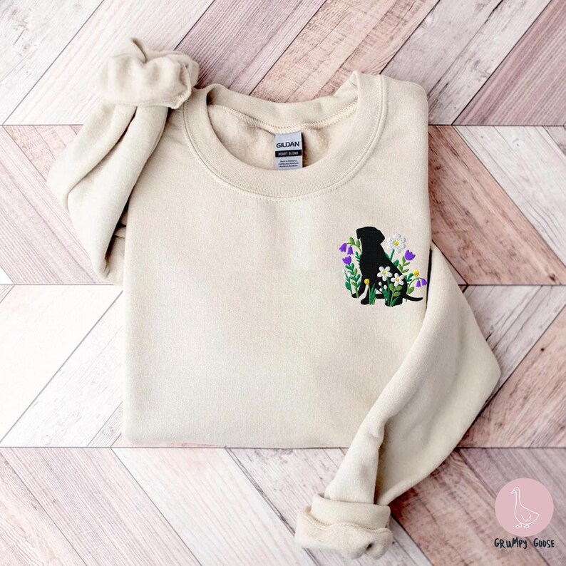 A sand sweatshirt featuring an embroidered black Lab dog sitting amidst a charming patch of wildflowers, adding a touch of nature and whimsy.