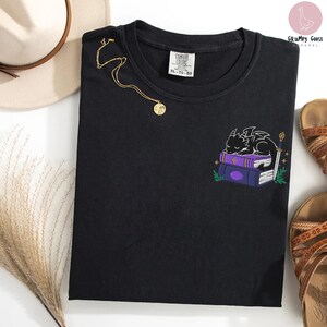 May include: Black crew neck t-shirt with an embroidered design of a sleeping dragon on purple books, a sword, and greenery. A gold necklace with a pendant is draped on the shirt. The shirt has a Comfort Colors tag.