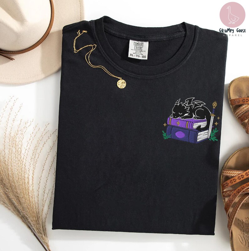 May include: Black crew neck t-shirt with an embroidered design of a sleeping dragon on purple books, a sword, and greenery. A gold necklace with a pendant is draped on the shirt. The shirt has a Comfort Colors tag.