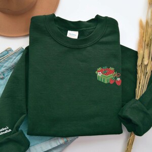 May include: Dark green sweatshirt featuring an embroidered strawberry basket design. The design includes red strawberries, white flowers, and green leaves. The sweatshirt is made of a soft material and has a crew neck.
