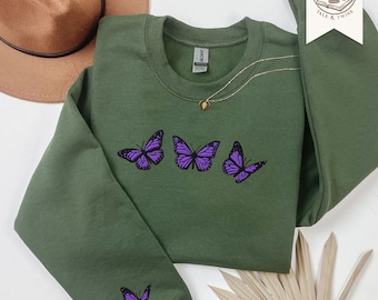 May include: An olive green crewneck sweatshirt featuring three embroidered purple butterflies across the chest and one additional purple butterfly embroidered on the lower left sleeve. The garment has a ribbed collar, cuffs, and hem.
