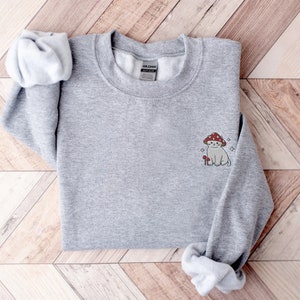 K&ouml;nnte beinhalten: Grauer Pullover mit einem kleinen gestickten Design eines wei&szlig;en Katers, der einen roten Pilz auf dem Kopf tr&auml;gt. Die Katze sitzt neben einem kleinen Pilz.