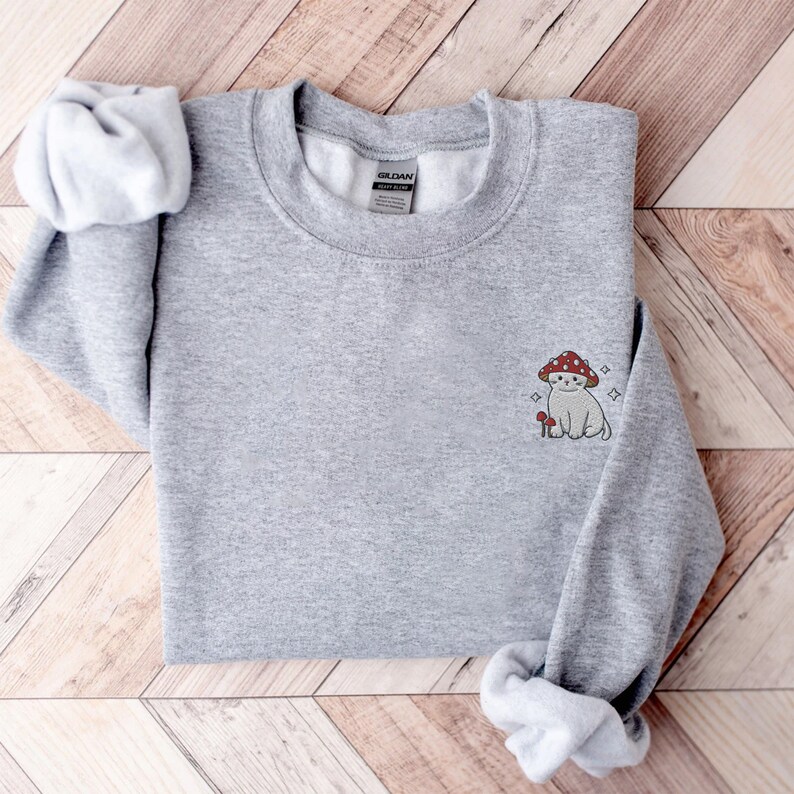 K&ouml;nnte beinhalten: Grauer Pullover mit einem kleinen gestickten Design eines wei&szlig;en Katers, der einen roten Pilz auf dem Kopf tr&auml;gt. Die Katze sitzt neben einem kleinen Pilz.