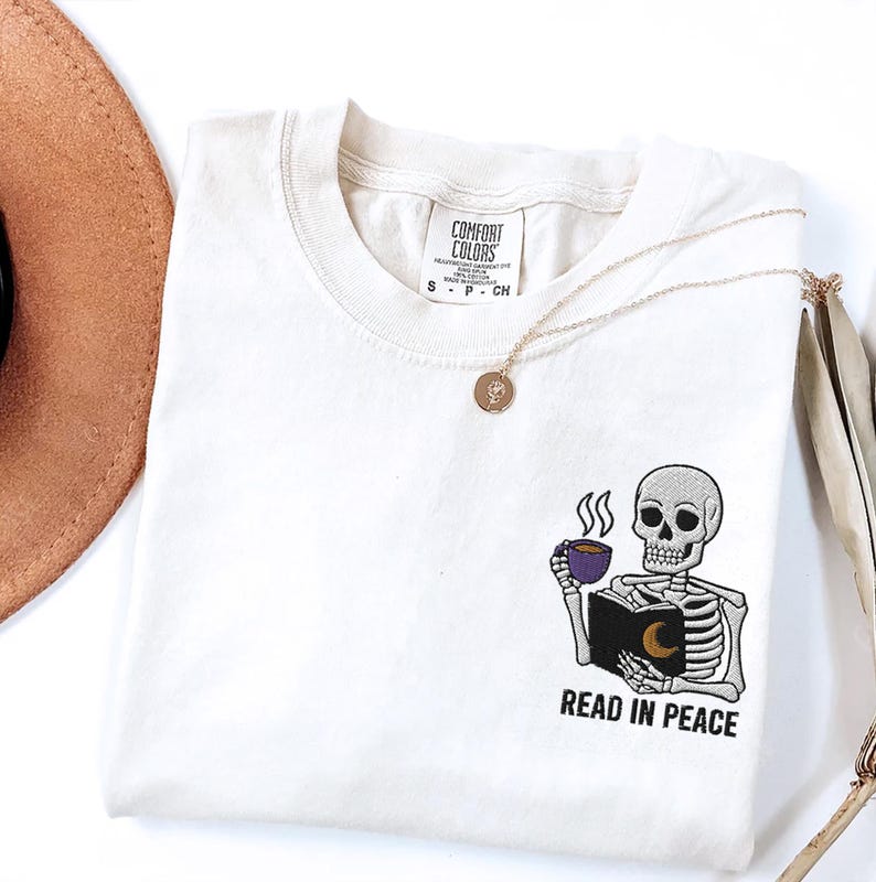 Puede incluir: Una camiseta blanca con un dise&ntilde;o bordado de un esqueleto sosteniendo un libro y una taza de caf&eacute;. El texto "Read in Peace" est&aacute; debajo del esqueleto.
