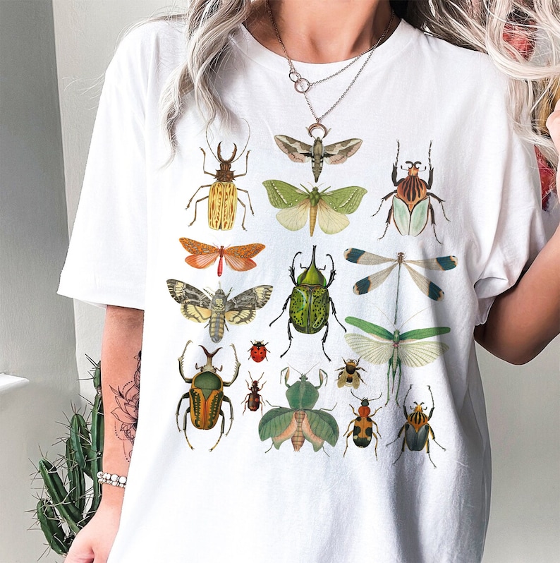 May include: White t-shirt with a colorful insect print. The insects include butterflies, beetles, and dragonflies. The shirt has a crew neck and short sleeves.