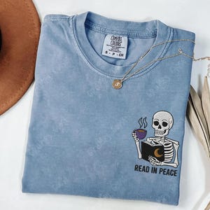 Puede incluir: Una camiseta azul claro con un esqueleto sosteniendo un libro y una taza de caf&eacute;. El texto "Read in Peace" est&aacute; bordado debajo del esqueleto.