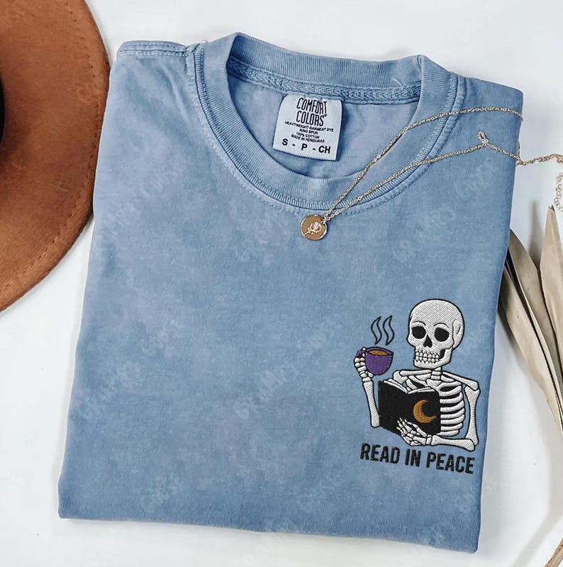 Puede incluir: Una camiseta azul claro con un esqueleto sosteniendo un libro y una taza de caf&eacute;. El texto "Read in Peace" est&aacute; bordado debajo del esqueleto.