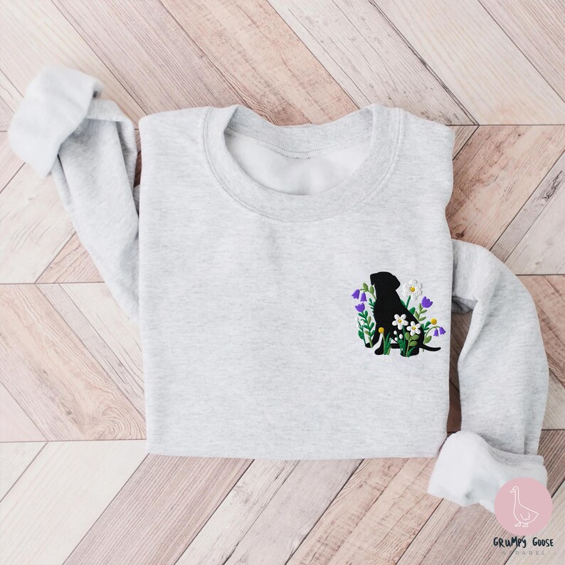 Pu&ograve; includere: Una felpa grigio chiaro con una silhouette nera di un cane circondata da fiori ricamati bianchi, viola e verdi. La felpa ha maniche lunghe con polsini arrotolati.