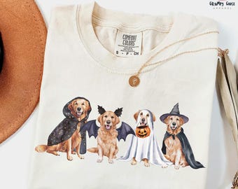 May include: A cream-colored t-shirt featuring a graphic of four golden retriever dogs dressed in Halloween costumes: a wizard with a star-patterned cape, a bat with black wings, a ghost holding a pumpkin bucket, and a witch in a black hat.
