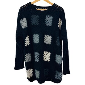 May include: A long-sleeve crochet sweater in dark navy blue featuring a repeating pattern of square motifs in shades of light blue, gray, and off-white. The garment has a round neckline and an open-knit texture throughout the body and sleeves.