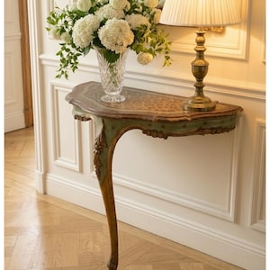Peut inclure: Une console murale demi-lune avec une finition peinte en vert clair et des accents de feuille d'or, un plateau à motifs bruns et un pied unique incurvé. La table présente des détails sculptés complexes et un bord festonné lisse.