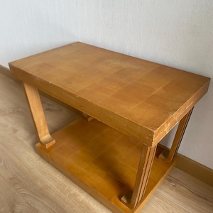 Peut inclure: Une table d'appoint rectangulaire en bois avec une finition brun clair et un motif de placage en damier visible sur le dessus. La table dispose d'une étagère inférieure et de quatre pieds incurvés reliant le plateau à la base, créant une structure à deux niveaux.