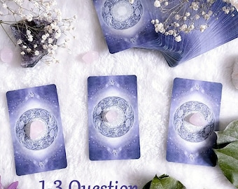 Può includere: Un'impostazione di lettura dei tarocchi con tre carte disposte, ciascuna con un design circolare e un cristallo rosa. Sono presenti anche un mazzo di carte e fiori decorativi. Il testo "1-3 Question Tarot Reading" è visualizzato.