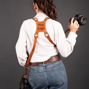May include: A brown leather camera harness with the text "Your Logo" on the chest plate. The harness is worn over a white shirt and blue jeans. A black camera is attached to the harness.