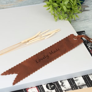 Peut inclure: Un marque-page en cuir marron avec les mots "Lovely Made" gravés dessus. Le marque-page est posé sur une pile de livres avec une couverture blanche.