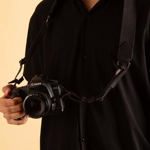 K&ouml;nnte beinhalten: Ein schwarzes Kameraband mit einem Metallring und eine schwarze Kamera mit einem Canon EOS-Objektiv.